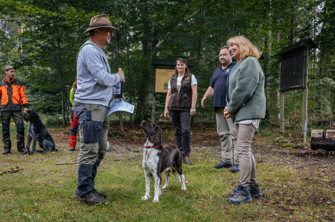 Petra Köpping beglückwünscht einen Hundehalter zur bestandenen Ausbildung, ein Hund steht zwischen beiden. Im Hintergrund stehen weitere Absolventen der Ausbildung mit Hund. 