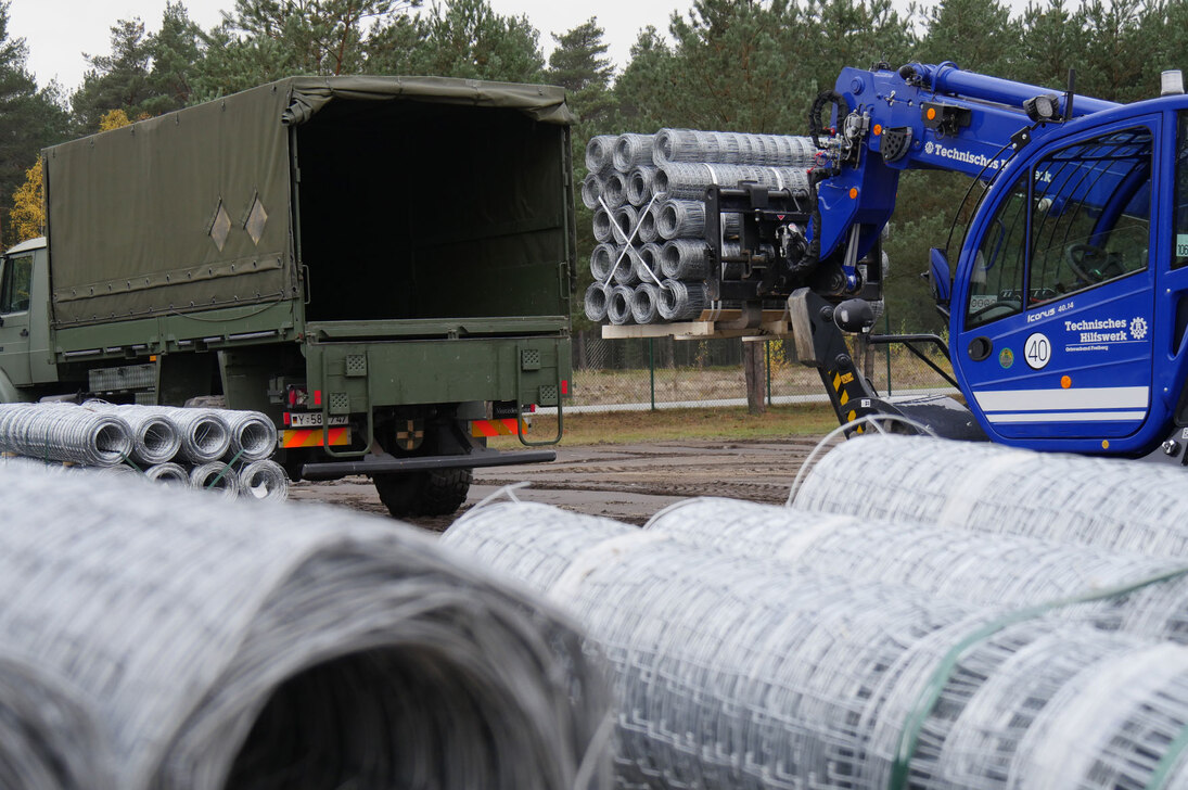 Ein LKW der Bundeswehr und ein Einsatzfahrzeug des Technischen Hilfswerkes stehen auf auf einem Lagerplatz unzähligen Rollen von Maschendraht